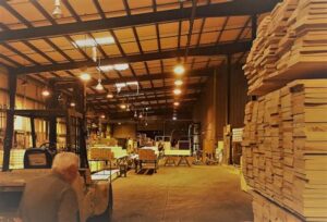Inside a lumber mill with stacks of cut wood, a forklift in operation, and workers handling materials under bright industrial lighting