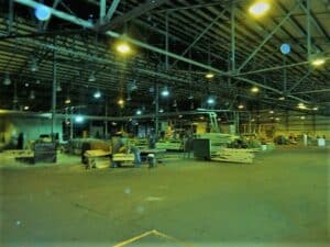 Interior of a lumber mill with workers operating equipment, stacks of cut wood, and industrial lighting illuminating the large open space