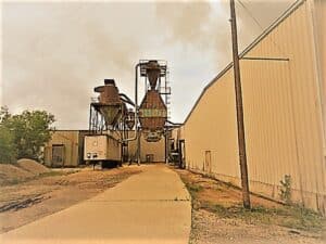 Industrial facility with large metal silos, pipes, and equipment between two long buildings, viewed from a concrete pathway