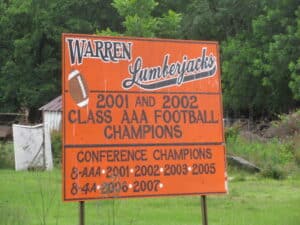 Orange signboard reading ‘Warren Lumberjacks, 2001 and 2002 Class AAA Football Champions, Conference Champions 2001-2007,’ placed outdoors with trees in the background