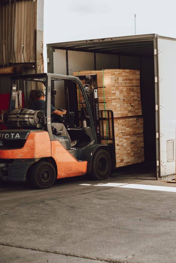 Forklift loading a large bundle of lumber into the back of a white delivery truck at a lumber facility.