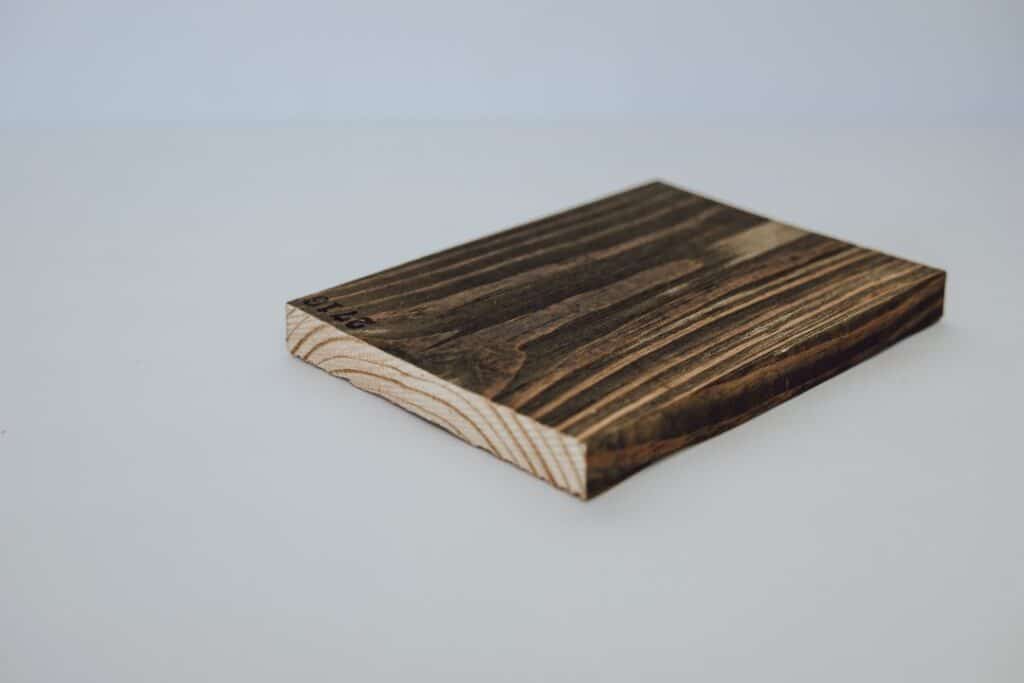 Single stained wooden board with visible grain patterns, lying flat on a white surface, showing its cut edge