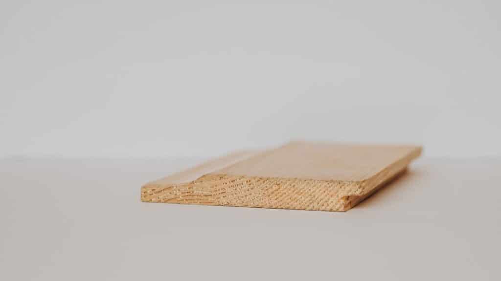 Close-up side view of a single unfinished shiplap wood board lying flat on a light surface, showing its rabbeted edge joint profile