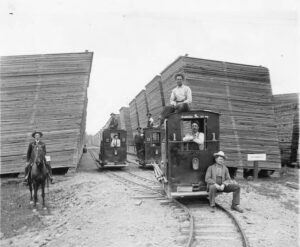Historical photo of workers and small locomotives in a lumber yard, with large stacks of lumber towering on both sides